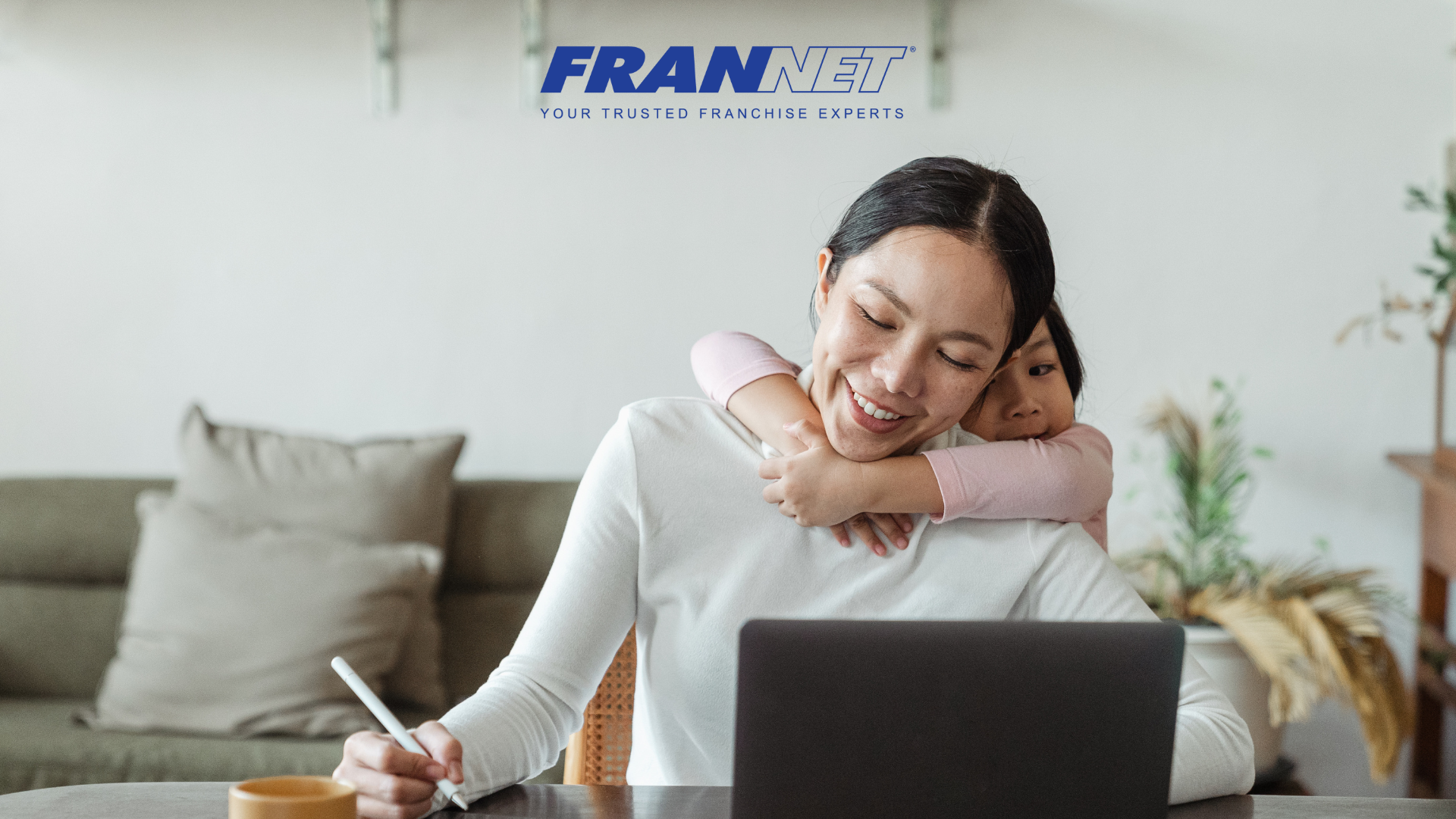 Mom working at computer with toddler hanging on her neck representing best franchises for moms